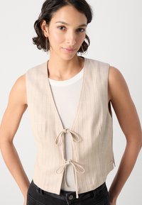 Beige mouwloze vest met verticale strepen, sluiting met voorste strik en een gladde textuur. Draag het over een witte geribde top, gecombineerd met zwarte spijkerbroek.