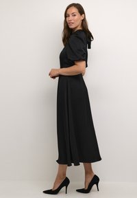 Rochie midi din satin negru, cu mâneci bufante, detaliu cu fundiță la spate și o siluetă fluidă. Asortată cu pantofi cu toc ascuțit negri.
