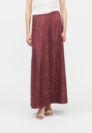 Mujer con falda larga de encaje burdeos y top blanco sin mangas, combinados con sandalias de tacón beige con punta abierta, posando frente a un fondo blanco.