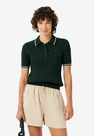 Femme aux cheveux courts et bouclés portant un polo en tricot vert foncé à manches courtes et un short beige à cordon, tenant un sac à main noir.