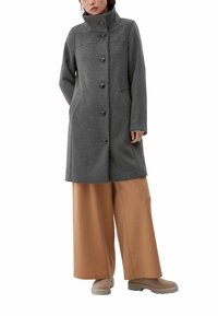 Grå uldfrakke med høj krave, seks knapper, sidelommer og et design i midterlængde, kombineret med brede beige bukser og let fodtøj.