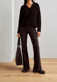 Persona con un maglione a coste nero e jeans neri a zampa d'elefante, in piedi su un pavimento in legno contro un muro semplice, mentre tiene una borsa a tracolla nera.