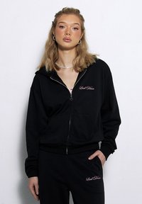 Czarna bluza z kapturem na zamek, wykonana z miękkiego materiału, z różowym logo na lewej piersi oraz ściągaczami przy rękawach i u dołu. Parę pasujących czarnych spodni.