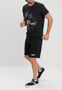 Camiseta negra con un gráfico de calavera y texto, combinada con pantalones cortos negros que presentan un logo blanco de "Creeks", y zapatillas negras con acentos blancos.