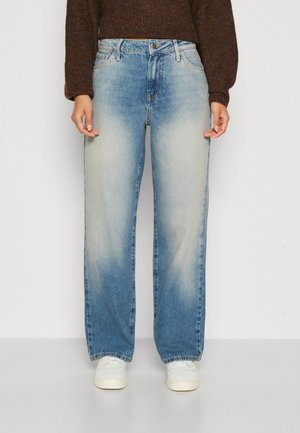 Femme portant un jean large délavé bleu clair, un pull marron à manches longues et des baskets blanches, debout devant un fond uni.