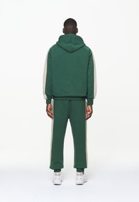 Felpa verde con cappuccio e pantaloni sportivi con strisce laterali beige; tessuto misto cotone; polsini e vita elastici; sneakers bianche. Sfondo neutro.
