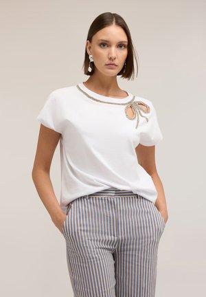 Frau trägt weißes kurzärmeliges Oberteil mit Strassschleifen-Detail nahe der Schulter und gestreifte blaue, weiße und braune Hose, Hände in den Taschen.