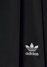 Czarna tkanina z teksturowanym splotem, na której znajduje się małe białe haftowane logo Adidas z trzema akcentami liści i trzema poziomymi paskami.
