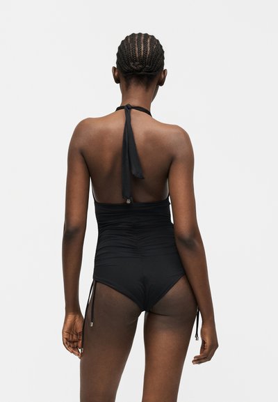 Γυναίκα με πλεξούδες, φορώντας μαύρο μαγιό με λαιμόκοψη halter, ποζάρει με την πλάτη προς το φακό πάνω σε απλό φωτεινό φόντο.