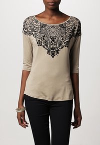 Beige long-sleeve t-shirt with black floral lace detailing across the shoulders. Soft fabric, relaxed fit, round neckline. Black pants worn.