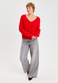 Un pull rouge oversize en tricot avec un décolleté en V profond, associé à un jean gris à jambe large et des chaussures noires à bout pointu.