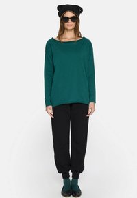 Długi rękaw, turkusowa bluzka z szerokim dekoltem, w zestawieniu z czarnymi spodniami dresowymi. Czarna dziana czapka z uszami kota i okulary przeciwsłoneczne uzupełniają stylizację.