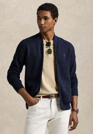 Jeune homme portant une veste zippée bleu marine, une chemise beige, un pantalon blanc, une ceinture marron et des lunettes de soleil accrochées au col de la chemise.