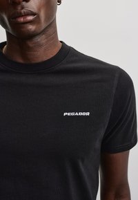 Person iført en enkel sort T-shirt med ordet Pegador i små hvide bogstaver på brystet, vist fra skuldre til midt på overkroppen.
