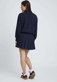 Sweatshirt bleu marine avec col et jupe plissée. Conçu avec une texture douce et associé à des baskets décontractées, présentant des accents blancs et bordeaux.