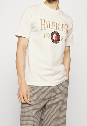 Homme portant un T-shirt blanc à manches courtes avec le texte et l'emblème "Hilfiger 1985 New York City", associé à un pantalon à carreaux.