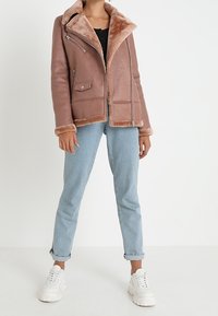 Giacca marrone con fodera in pelliccia sintetica, chiusura con zip davanti e due tasche laterali; presenta un colletto ampio e un tessuto texture; abbinata a jeans azzurri.