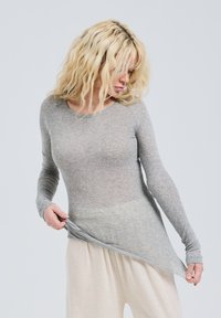 Maglione grigio a maniche lunghe con un corpo aderente e un orlo posteriore leggermente più lungo, realizzato in un tessuto trasparente e testurizzato. Abbinato a pantaloni beige chiaro.