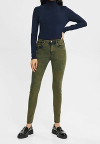 Jeans skinny verde oliva con un acabado texturizado, combinados con un cuello de tortuga ajustado de color azul oscuro y mocasines de plataforma negros.