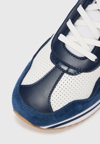 Marineblå og hvit atletisk sko med perforert skinn og semsket tekstur. Har hvite lisser og en kontrasterende gummisåle.