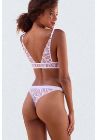 Femme aux cheveux bouclés portant un ensemble de lingerie en dentelle violette claire assortie, vue de dos sur un fond uni clair.