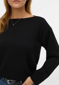 Melna garām piedurknēm topi ar laivas formas izgriezumu, mīkstu tekstūru un vaļēju piegriezumu. Ar nolaižamām pleciem un ribotām aprocēm. Kombinēts ar džinsu materiālu.