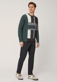 Maglione verde a maglia con blocchi di colore geometrici in bianco, grigio e marrone, abbinato a pantaloni scuri e sneakers con dettagli beige e neri.