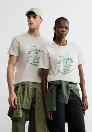 Dwie modelki w beżowych koszulkach Tommy's Gym Los Angeles, z zielonymi koszulami przewiązanymi w talii, pozujące na jednolitym tle.