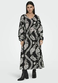 Pitkä musta mekko, jossa on valkoisia lehtikuvioita, V-pääntie, pitkät hihat ja vyötärölle kiinnitettävä vyö. Mallilla on päällä mustat nilkkurit.