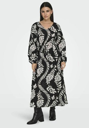 Langes schwarzes Kleid mit weißen blättrigen Mustern, mit V-Ausschnitt, langen Ärmeln und tailliertem Gürtel. Das Model trägt schwarze Knöchelstiefel.