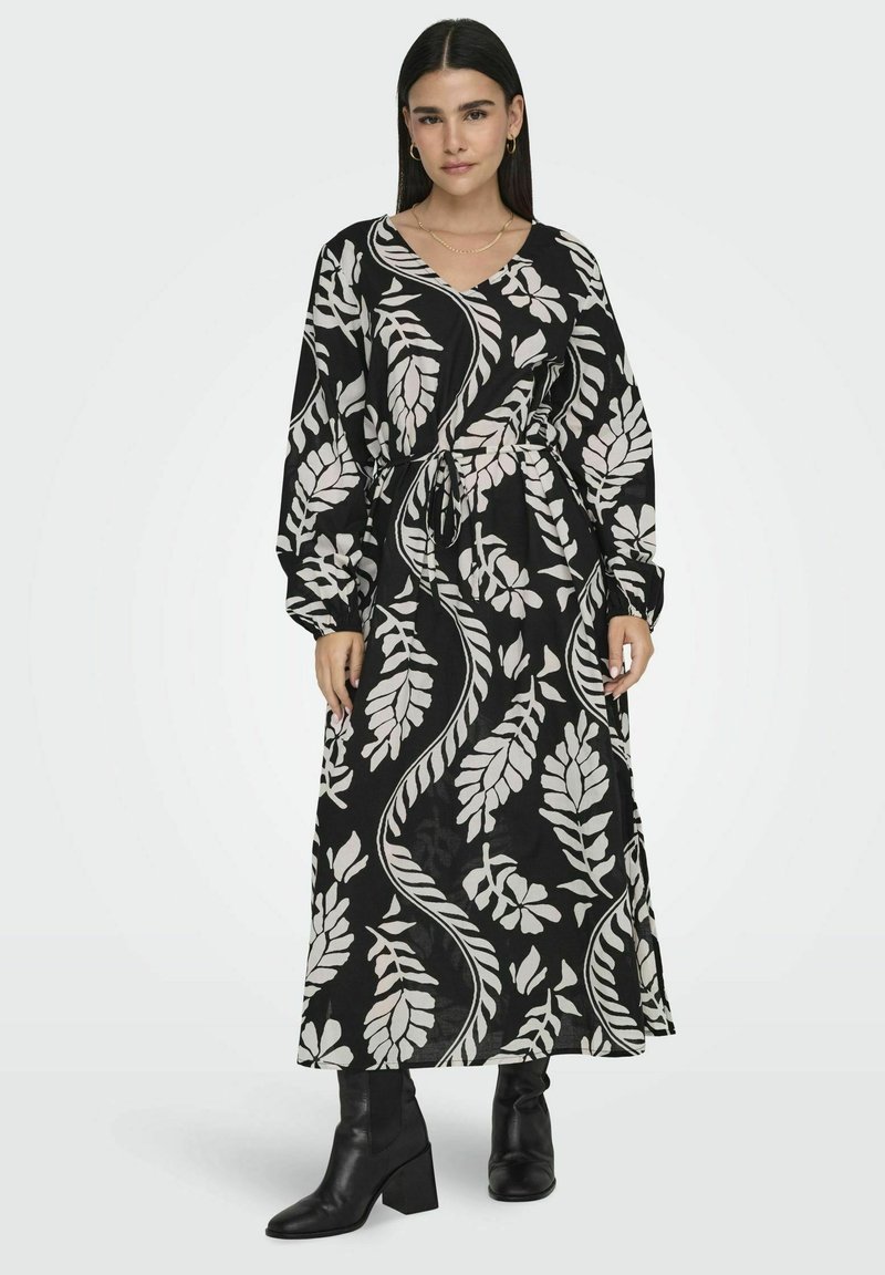 Pitkä musta mekko, jossa on valkoisia lehtikuvioita, V-pääntie, pitkät hihat ja vyötärölle kiinnitettävä vyö. Mallilla on päällä mustat nilkkurit.