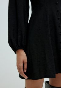 Schwarzes Kleid aus strukturiertem Stoff, lange gepuffte Ärmel, Knopfdetails und ein leicht ausgestellter Saum bis zur Mitte des Oberschenkels.