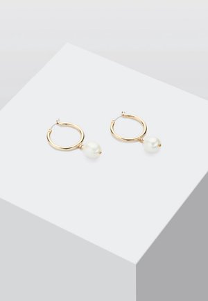 Boucles d'oreilles créoles en or avec une perle blanche suspendue à chacune, présentées sur une surface blanche inclinée.