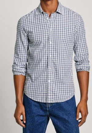 Hombre vistiendo una camisa de cuadros azul y blanca de manga larga con las mangas remangadas y jeans de mezclilla azules, de pie frente a un fondo liso.