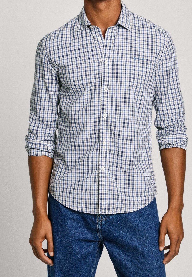 Uomo che indossa una camicia a maniche lunghe a quadri blu e bianchi con le maniche arrotolate e jeans di denim blu, in piedi contro uno sfondo neutro.