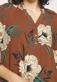 Top in tessuto marrone con scollatura a V, caratterizzato da grandi motivi floreali crema e verde, e tre bottoni al collo.