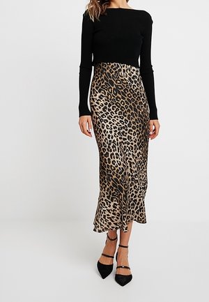 Kobieta ubrana w czarną bluzkę z długim rękawem, midi spódnicę w cętki lamparta oraz czarne szpilki z paskami wokół kostek, stojąca na jednolitym tle.