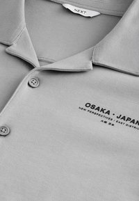 Siva polo majica napravljena od mekog materijala, sa klasičnim ovratnikom i dva dugmeta. Na prsima je otisnut tekst "OSAKA • JAPAN."