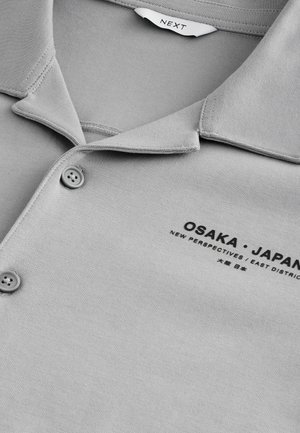 Šedé polo tričko vyrobené z měkké tkaniny, s klasickým límcem a dvěma knoflíky. Na hrudi je vytištěn text "OSAKA • JAPONSKO."