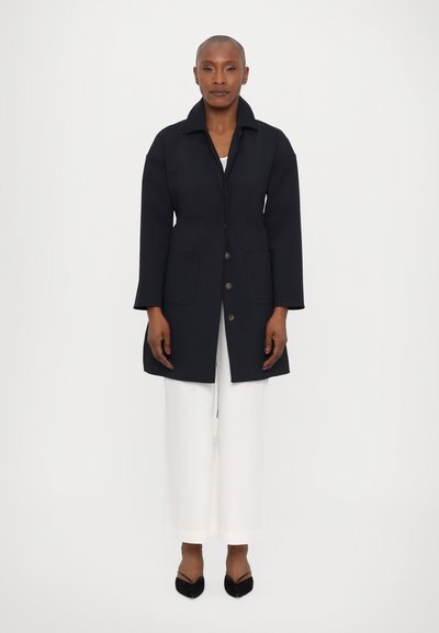 Σακάκι από ναυτικό μαλλί με καπιτονέ λαιμό, κλείσιμο με κουμπί και δύο μπροστινές τσέπες. Το σακάκι έχει ευθεία σιλουέτα και μακριά μανίκια.