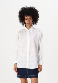 White button-up shirt with tonal "HUGO" logo print, long sleeves, and a classic collar, paired with a dark denim skirt.