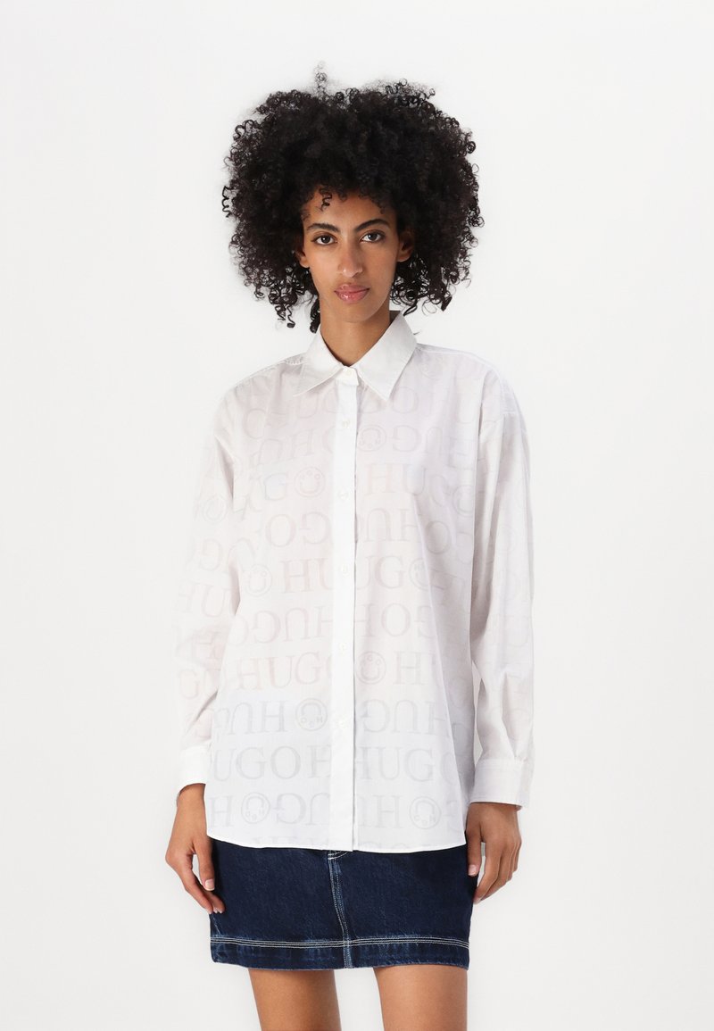 White button-up shirt with tonal "HUGO" logo print, long sleeves, and a classic collar, paired with a dark denim skirt.
