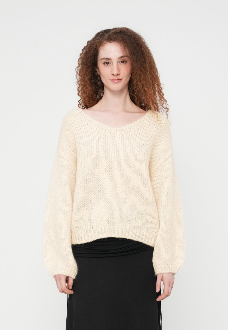 Ελαφρύ εκρού πλεκτό πουλόβερ σε μεγάλο μέγεθος με V-λαιμόκοψη, ριμπ τελείωμα και φαρδιά μανίκια. Μαλακή υφή, άνετη εφαρμογή, σε συνδυασμό με μαύρη φούστα.