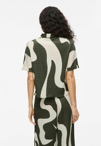 Short-sleeved cropped shirt in green and beige abstract pattern, featuring a textured fabric and rolled cuffs. Back view shows a relaxed fit.