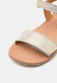 Metallisk guld sandalett med ett brett band och en mjuk, tanfärgad sula. Platt sula med subtil textur, med en lätt sliten finish.