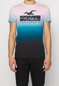 Camiseta de hombre con un degradado de rosa claro a verde azulado oscuro, con un logo gráfico negro y texto que dice "Hollister California". Mangas cortas.