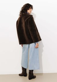 Manteau en fausse fourrure marron avec un large col et des poches latérales, associé à un jean bleu clair à jambes larges et des bottes hautes marron foncé.