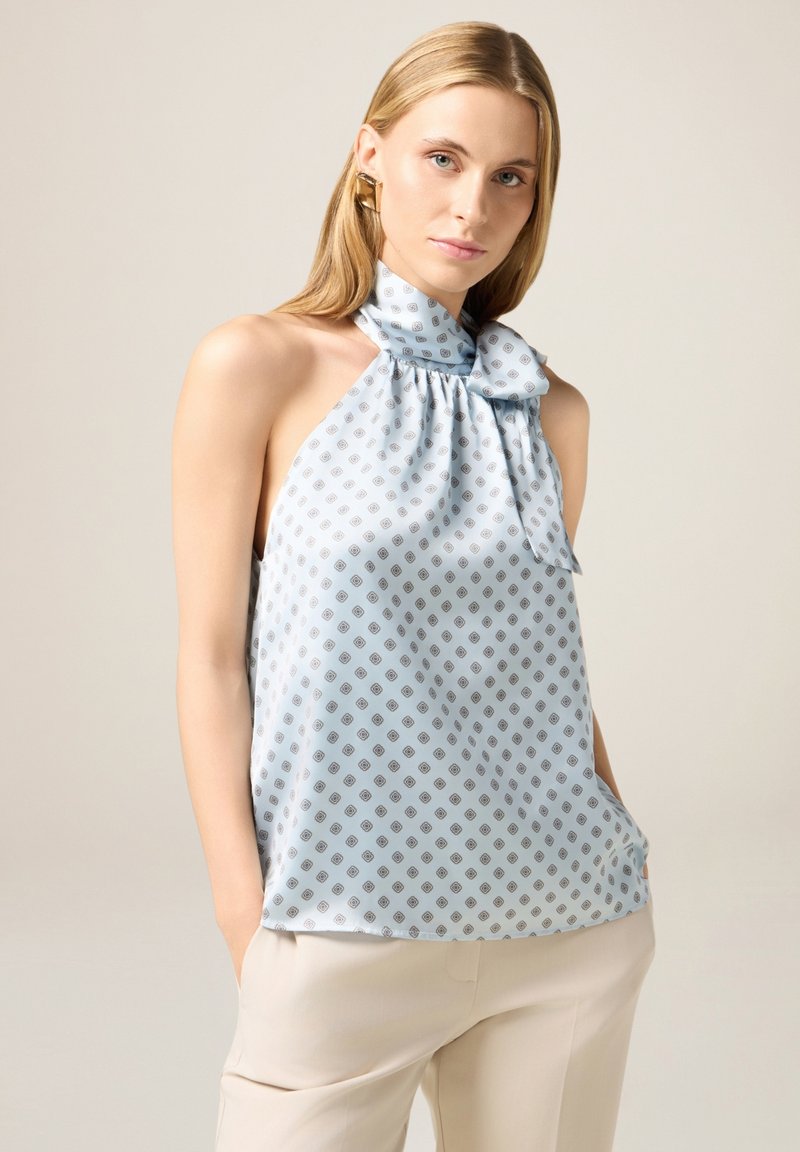 Top halter azzurro chiaro realizzato in tessuto liscio, con piccoli motivi a diamante e un dettaglio a fiocco al collo. Abbinato a pantaloni chiari.