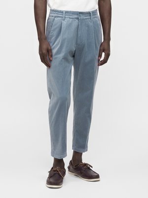 Hombre con pantalones de pana azul claro y zapatos náuticos de cuero marrón oscuro, de pie frente a un fondo blanco simple.