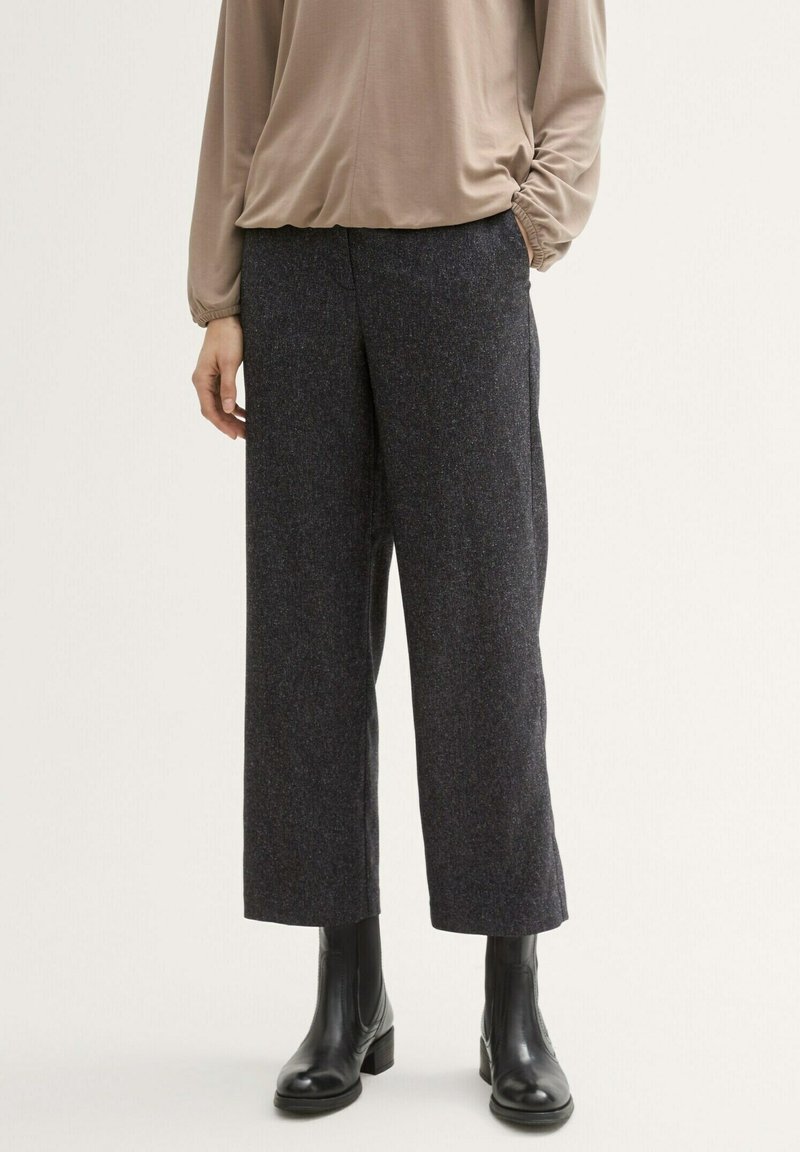 Personne portant un pantalon large court de couleur sombre, un haut beige à manches longues et des bottines noires sur un fond uni.